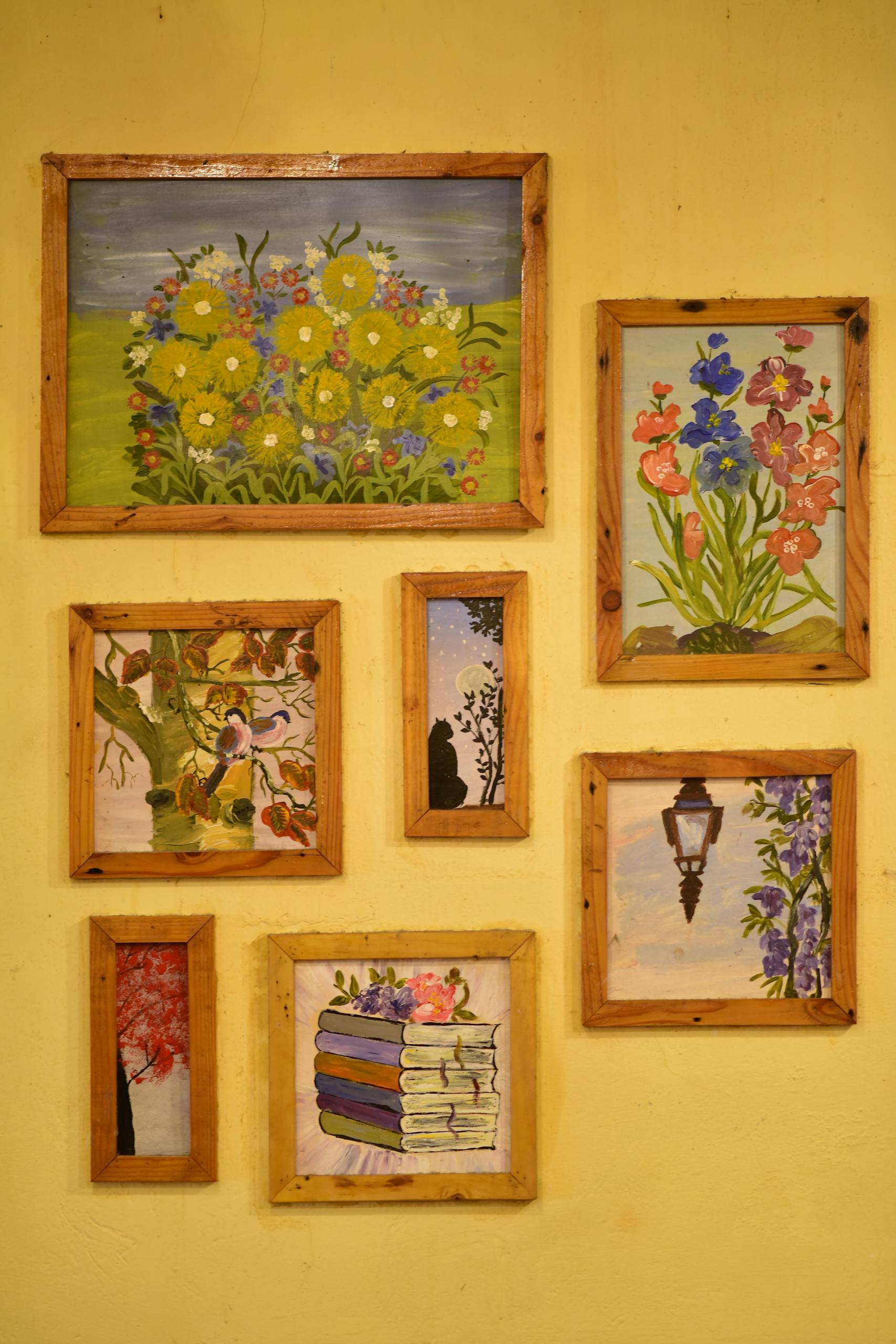 A set of vibrant framed paintings displayed on a yellow wall, featuring flowers and books.