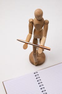 A wooden mannequin holds a pen above a spiral-bound notebook, ideal for creativity concepts.
